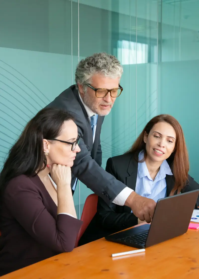 three professionals with obscured faces at a meeting, one standing and two seated with a laptop
