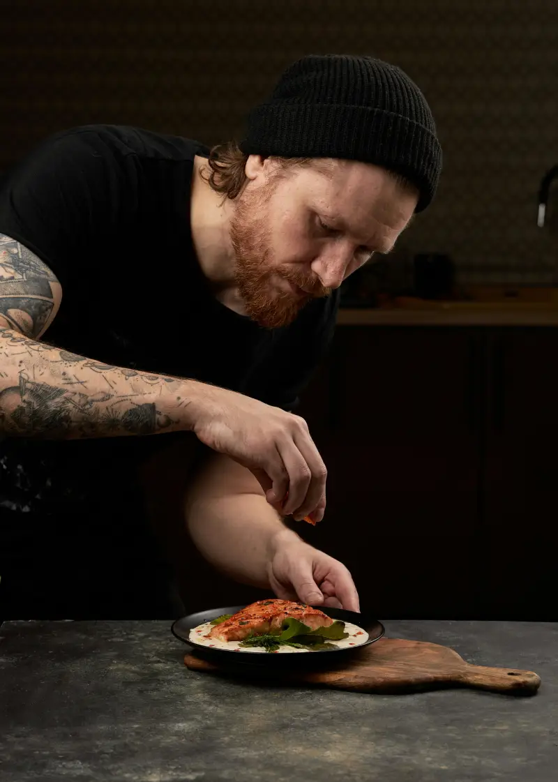 A person in a black beanie prepares a plated dish of salmon with herbs, enhancing it with a sprinkle of seasoning A person in a black beanie prepares a plated dish of salmon with herbs, enhancing it with a sprinkle of seasoning