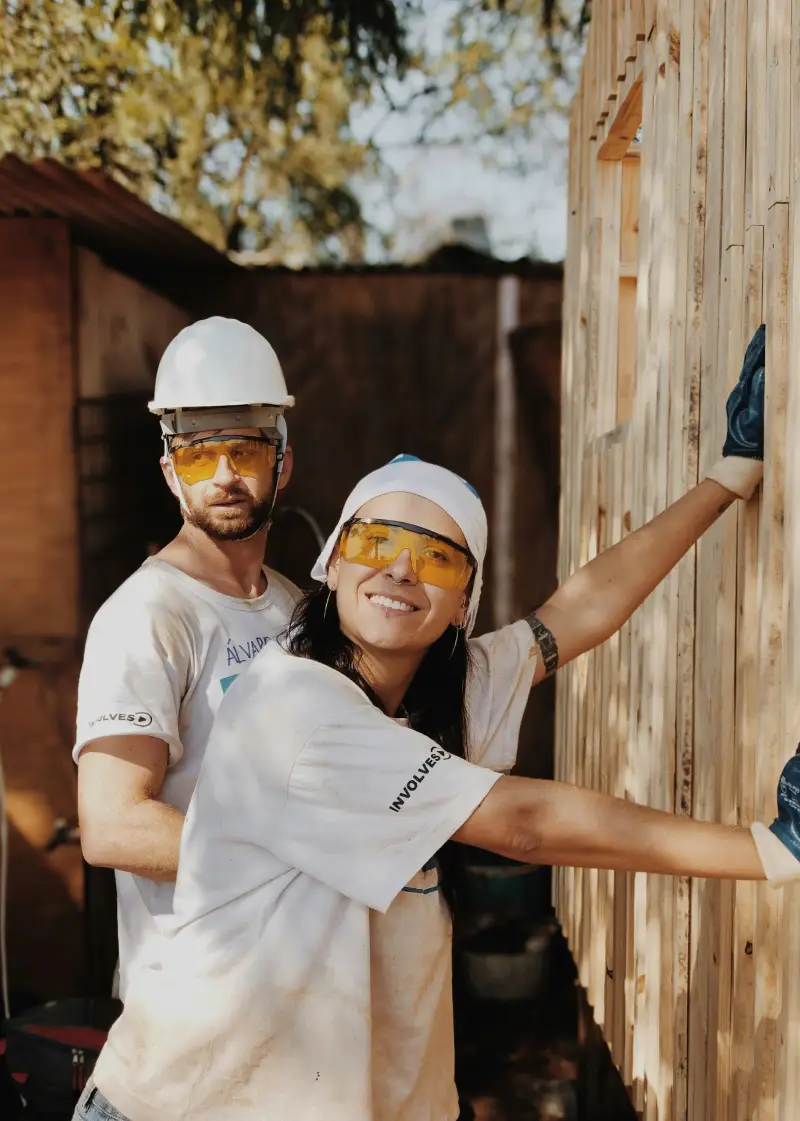 Two construction workers in hard hats and gloves collaborate on a wooden wall in a sunny outdoor setting. Tools are visible nearby